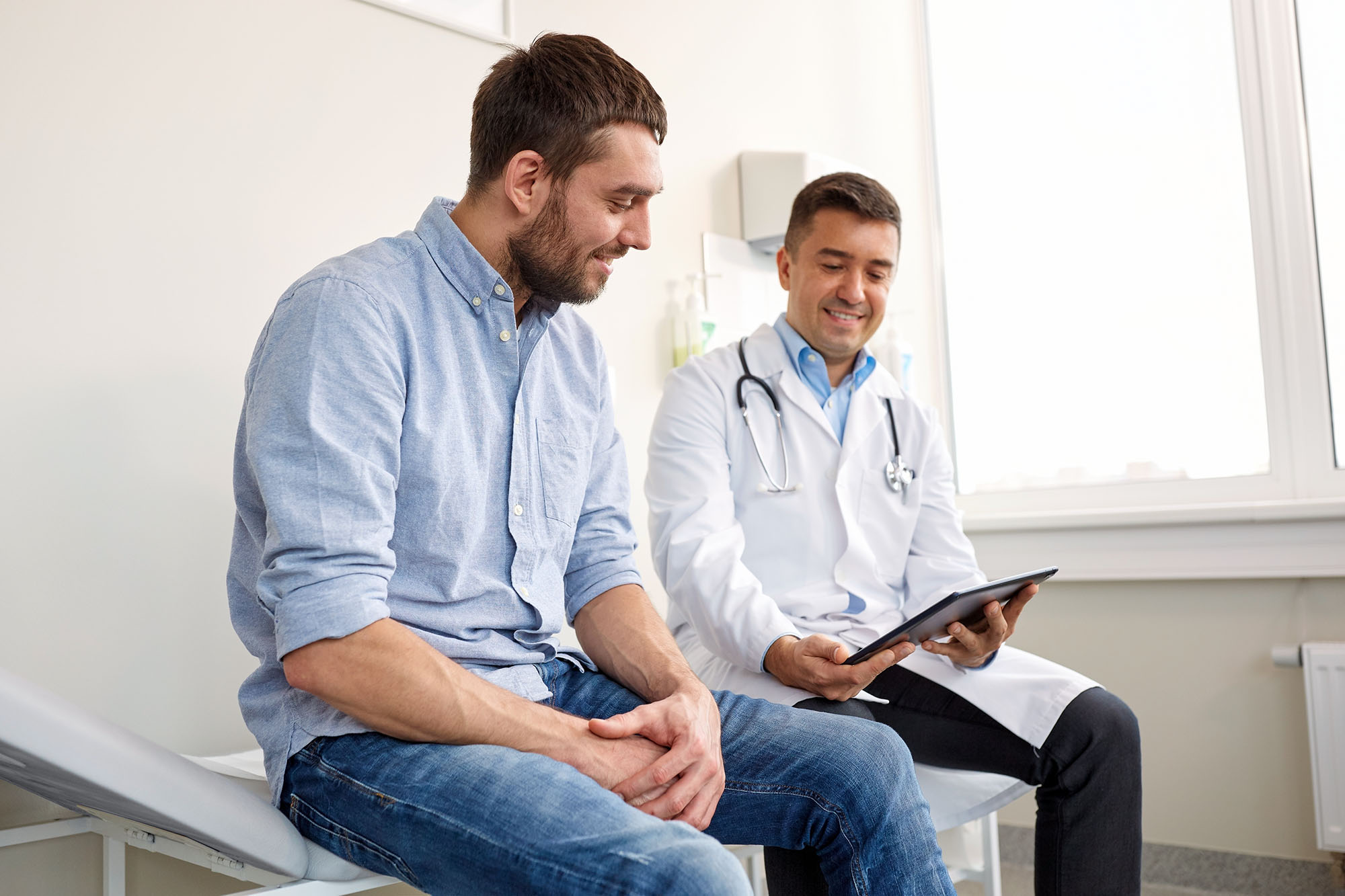 medicine-healthcare-people-concept-smiling-doctor-with-tablet-pc-computer-young-man-patient-meeting-hospital doctor with tablet computer speaking with young man patient in exam room